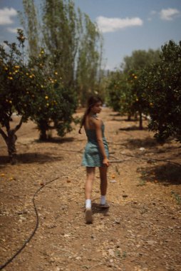 Güneşli bir günde, güzel ağaçlarla çevrili bir meyve bahçesinde yürüyen genç bir kız huzurlu bir atmosfer yaratıyor.