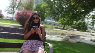 Genç bir kadın, güneşli bir günde, yeşil bir günde, bir bankta, akıllı telefon kullanıyor.