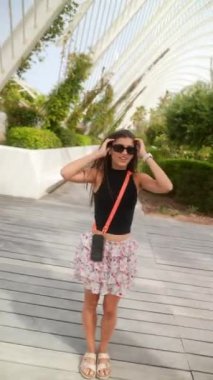 Güneş gözlüğü takan şık bir genç kadın, siyah bir bluz ve çiçekli bir etek açık havada poz veriyor.