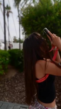 Uzun saçlı genç bir kadın telefonuyla lüks ve manzaralı bir parkta güzel anları yakalıyor.