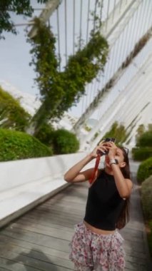 Bir kadın, uzun yeşil bitkiler ve palmiye ağaçları olan güzel, yemyeşil bir şehir bahçesinde anları yakalamak için kamera kullanır..