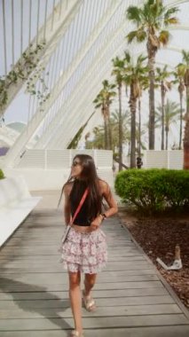 Güneş gözlüğü takan ve çiçekli etek giyen şık genç bir kadın, palmiye bahçesinde yürürken güneşli bir günün tadını çıkarıyor..