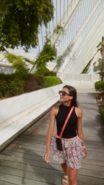 Çiçekli etekli ve siyah bluzlu bir kadın modern serada tropik bitkilerle yürüyor, güneş gözlüğü takıyor..