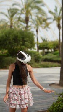 Genç bir kadın dışarıda sanal gerçeklik deneyiminden zevk alır. VR kulaklık takıyor ve güneşli bir parkta duruyor..