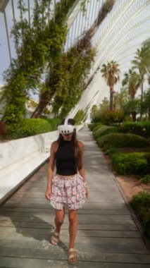 Genç bir kadın, beyaz bir miğfer ve siyah bir bluzla modern bir bahçe yolunda yürüyor..