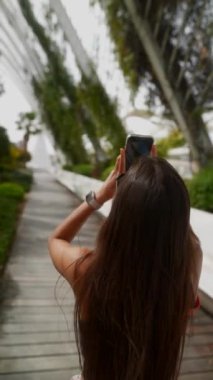 Bir kadın, modern ve yemyeşil bir açık hava ortamında fotoğraf çekiyor, manzaranın tadını çıkarıyor.