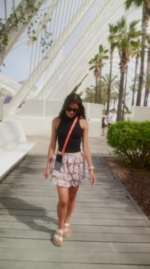 Siyah bluzlu, çiçekli etekli, kendine güveni tam bir kadın parkta modern bir mimari yapının altında yürüyor..