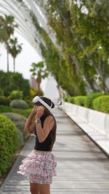 Çiçekli etekli genç bir kadın yeşil ve beyaz yapıları olan manzaralı bir ortamda VR kulaklık kullanıyor..