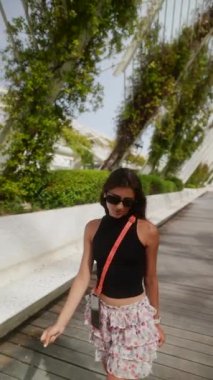 Siyah bluzlu ve çiçekli bir etek giyen genç bir kadın güneşli bir günde modern bir şehir bahçesinde keyifle yürüyor.