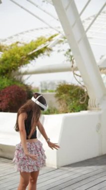 VR kulaklık takan genç bir kadın yeşillik ve modern mimari ile açık hava ortamını keşfediyor..
