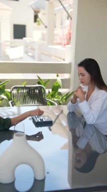 İki kadın bir satranç oyununa odaklanmış, açık havada yansıtıcı bir masada, stratejik düşüncenin derinliklerinde..