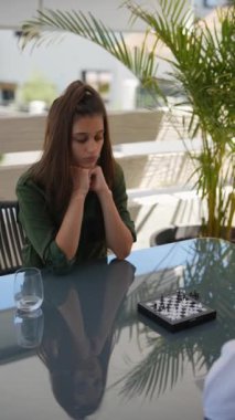 İki genç kadın dışarıda cam bir masada satranç oynuyorlar. Odaklandıkları ve stratejik düşüncelerini sergiliyorlar.