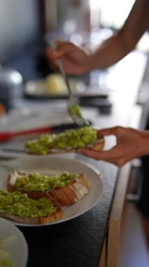 Avokado ezmesini modern bir mutfakta, arka planda bulanıklık olan ekmek dilimlerine saçan ellerin yakın çekimi..