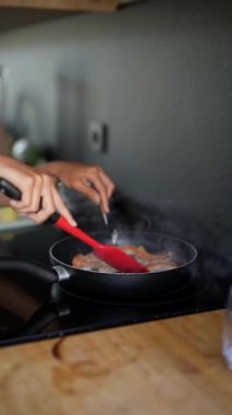 Elleri kızartma tavasında domuz pastırması pişirirken fırının üstünde kırmızı bir spatula kullanılıyor..