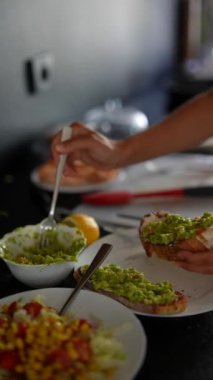 Modern bir mutfakta, bir elin tostun üzerine çeşitli taze malzemelerle avokado sürdüğü görülür.