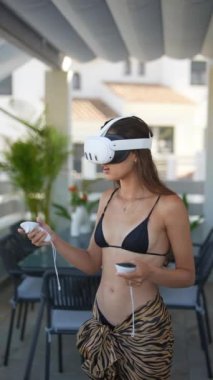 VR kulaklık ve kumanda takan, terasta mayo giymiş, tropikal dekorlu genç bir kadın..