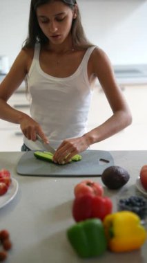 Modern bir mutfakta, bir kadın salatalıkları domates, biber ve avokado gibi taze ürünlerle dilimler.