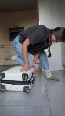 Modern mutfağında çırpınan bir kadın, ağzına kadar dolu bir bavulu kapatmakta zorlanıyor.