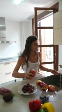 Beyaz kolsuz tişörtlü bir kadın yaratıcı bir şekilde mutfak tezgahına taze meyve ve sebze koyuyor.