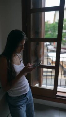 Rahat bir şekilde giyinen genç bir kadın, rahat bir iç mekandaki pencerenin yanındaki akıllı telefonuna odaklanmış durumda.