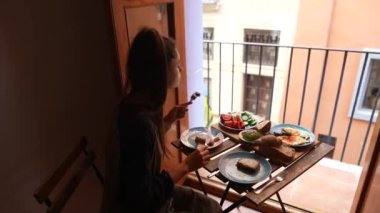 Bir kadın güneşli bir balkonda lezzetli bir geç kahvaltı yapıyor. Ekmeğin, taze meyvelerin ve sebzelerin tadını çıkarıyor.