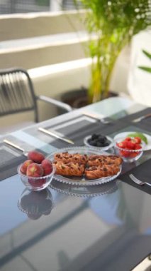 Bitkilerle dolu cam bir masada çörek, şeftali, çilek ve böğürtlenli açık hava kahvaltısı..