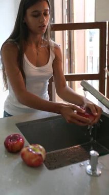 Genç kadın, çağdaş bir mutfakta doğal ışıkla çevrili akan suyla taze elmaları yıkıyor.