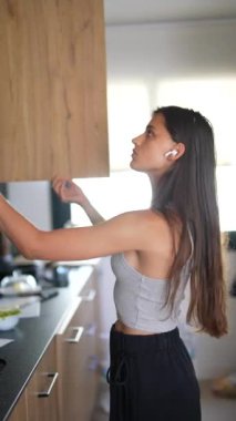 Genç bir kadın modern bir evde mutfak dolapları düzenliyor, kablosuz kulaklık takıyor, işine odaklanıyor.