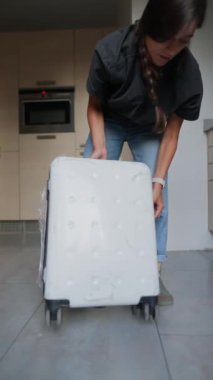 Bir kadın çömelmiş, modern bir mutfakta beyaz bir bavul hazırlıyor, seyahate ya da tatile hazırlanıyor.