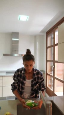 Genç bir kadın, çağdaş bir mutfakta bol ışık ve parlak bir tasarım ile sebzeleri dilimlerken görülüyor.