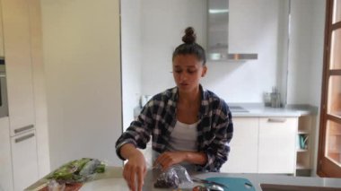 Ekose gömlekli genç bir kadın taze sebzelerle dolu modern bir mutfakta yemek pişiriyor ve rahat bir ortam yaratıyor.