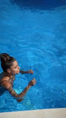 Bir kadın parlak güneş ışığı altında berrak bir mavi havuzda yüzer. Huzurlu bir ortamda rahatlık ve canlılık saçar.