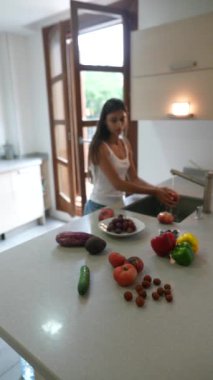 Modern bir mutfakta sebze yıkayan bir kadın, bir dizi taze ürün ve malzemeyle çevrili..