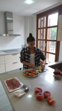 Genç bir kadın, taze meyve ve sebzelerle çevrili bir gülümsemeyle modern bir mutfakta sebze doğruyor..