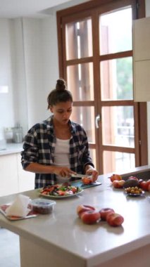 Modern bir mutfakta salata için domates doğrayan bir kadın. Etrafı taze malzemeler ve doğal ışıkla çevrili..
