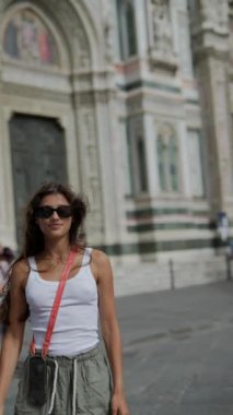 Tarihi binanın önünde poz veren şık bir kadın, güneş gözlüğü takıyor ve bir çanta tutuyor.