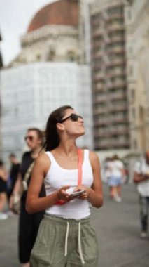 Güneş gözlüklü bir kadın tarihi bir şehri geziyor, gülümsüyor, akıllı bir telefon tutuyor, yaz seyahatlerinin tadını çıkarıyor.