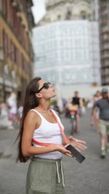 Yoğun bir şehirde güneş gözlüklü genç bir kadın, şehir manzarasını tesadüfen keşfeden akıllı bir telefonla şaşırmış görünüyor.