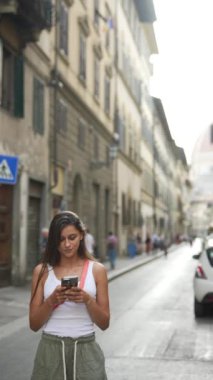 Arkasında binalar ve arabalar olan tarihi bir caddede genç bir kadın akıllı telefonunu kullanıyor.