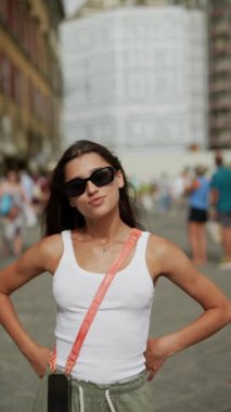 Güneş gözlüklü ve kolsuz tişörtlü şık bir kadın şehirde poz veriyor, modern şehir modasını ve özgüvenini simgeliyor.