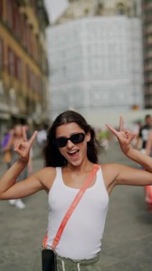 A woman outdoors, pointing at herself, in sunglasses and a white tank top, is enjoying a carefree moment in the city