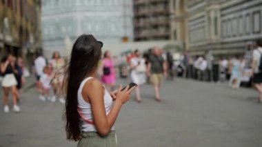 Akıllı telefon kullanan şık bir kadın hareketli bir şehir caddesinde yürüyor, modern şehir yaşam tarzını somutlaştırıyor.