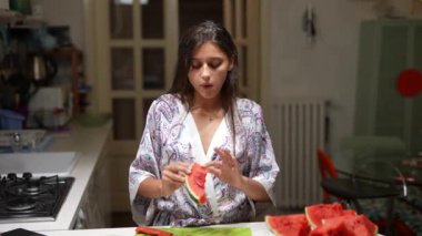 Genç bir kadın, daha fazla karpuz parçasıyla çevrili, modern bir mutfak ortamında bir dilim karpuzun tadını çıkarıyor..