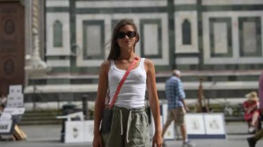 Günlük kıyafetli ve güneş gözlüklü bir kadın tarihi geçmişi olan bir caddede güven içinde poz veriyor..