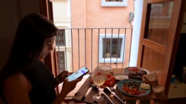 Sıcak bir şehir dairesinde, bir kadın kahvaltı ederken, akıllı telefonuna dalmış, şehir manzarasının tadını çıkarırken rahatlar.