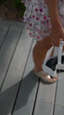 Çiçekli bir elbise giyen şık bir kadın güneşli bir günde elinde kamerayla ahşap bir yolda yürüyor.