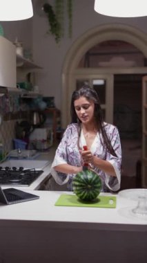 Modern bir mutfakta sakince karpuz doğrayan bir kadın dizüstü bilgisayar izliyor ve sakin bir atmosfer yaratıyor.