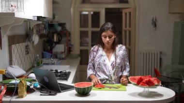 Genç bir kadın modern mutfakta karpuz dilimliyor ve eğleniyor, dizüstü bilgisayar tezgahta, rahat atmosfer