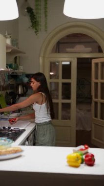 Çağdaş bir mutfakta, uzun siyah saçlı bir kadın tezgahta taze sebzelerin arasında yemek pişiriyor.