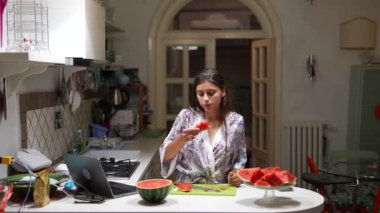 Modern bir mutfakta, bir kadın masada meyve ve dizüstü bilgisayarla karpuz keser ve rahat bir ev ortamını yaratır.
